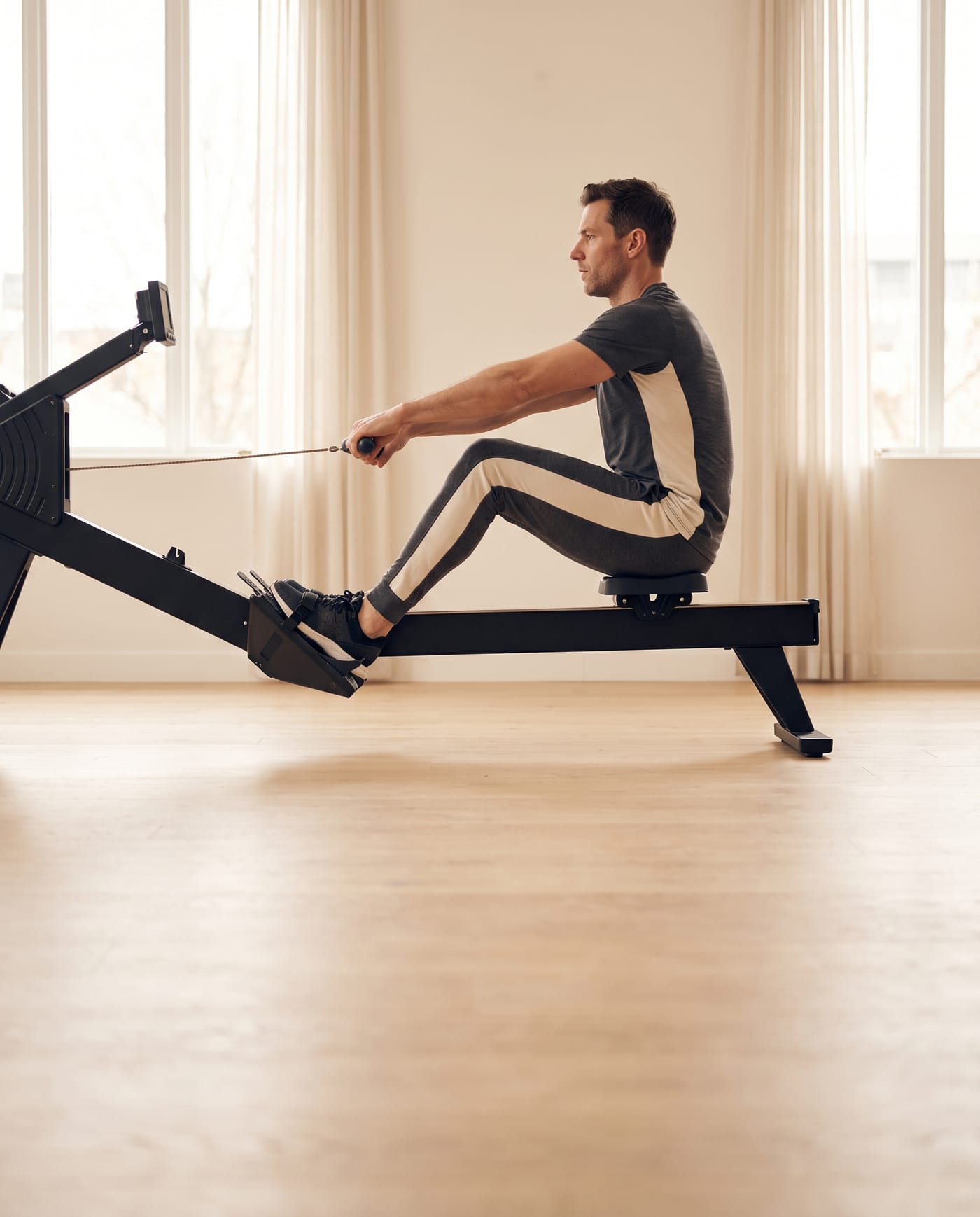 An athlete mid-stroke on a rowing machine beside tall windows